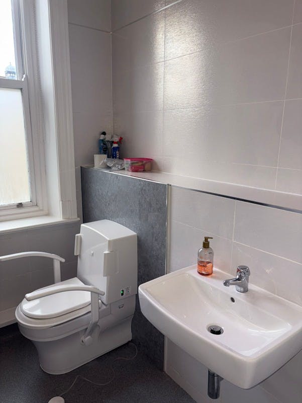 Accessible bathroom featuring a Closomat wash and dry WC with support arms and a wall-hung basin, finished with grey wall boards, off-white tiles and anti-slip vinyl flooring.